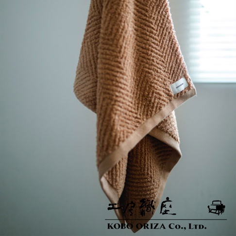 一條棕褐色人字紋有機棉浴巾，觸感柔軟，邊緣有米色滾邊，角落處縫有白色品牌標籤。