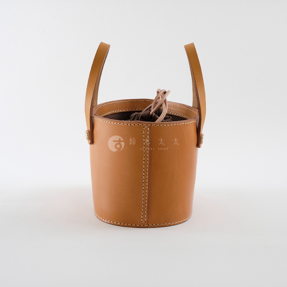 職人手作皮革束口手提包，採用天然皮革材質，呈現溫潤的淺棕色，搭配米白縫線。水桶造型，頂部有束口設計，附有兩條簡約的提把。