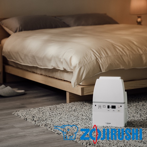 白色多功能智慧烘乾機置於臥室地板的毛毯上，旁邊是木製床架和米白色床單，床頭有兩個灰色枕頭。