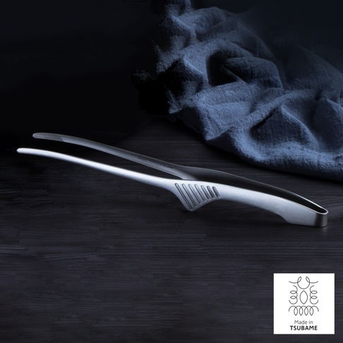 銀色金屬製不沾桌公筷夾/餐夾，一體成形設計，夾頭帶有防滑紋路，握柄處有扇形凹槽，置於深色木紋桌面上，背景為藍色皺褶布料。