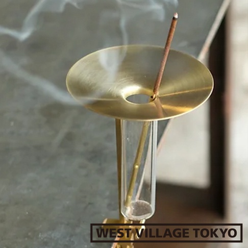 黃銅製的圓形線香盤，中央有圓孔，插入一根燃燒中的線香，盤下方為透明玻璃管，整體呈現金色光澤，有少量煙霧繚繞。