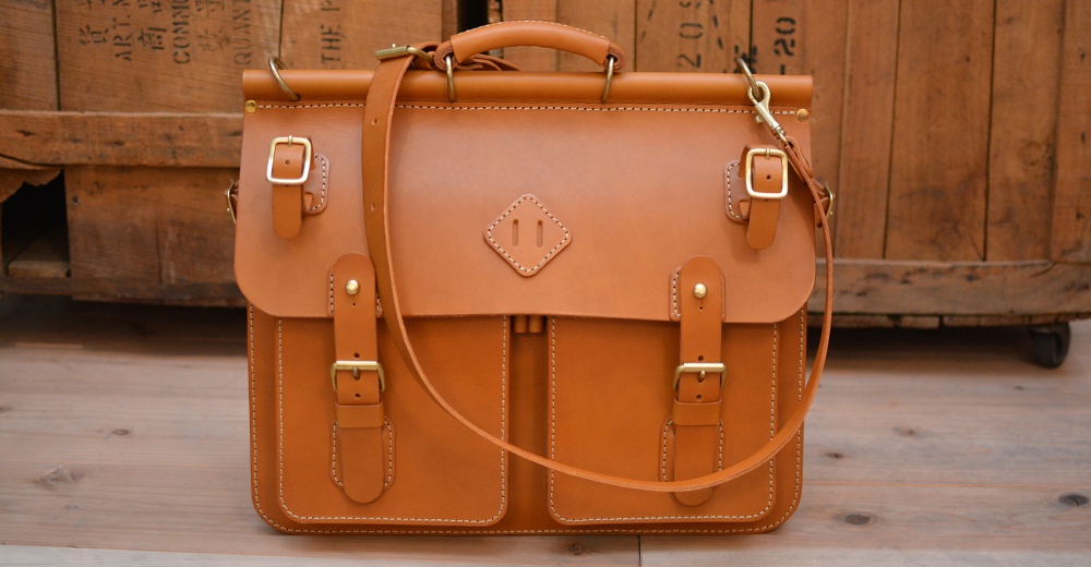 職人手作皮革旅行包 A-102，採用棕褐色光滑皮革製成，配有黃銅色搭扣和肩帶，正面有兩個翻蓋口袋和一個品牌標誌。
