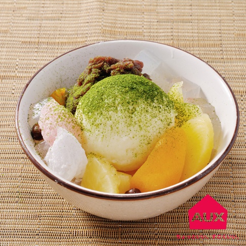 白色冰淇淋上撒滿翠綠抹茶粉，搭配紅豆泥、粉色冰淇淋、白色冰塊、果凍及黃色水果丁，盛裝於白色邊緣帶有褐色線條的碗中。