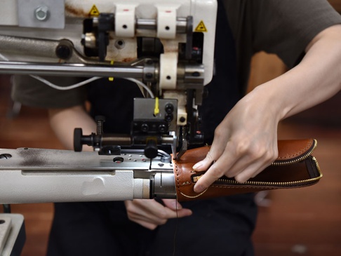 職人正使用工業縫紉機縫製一個棕色皮革製的筆袋，筆袋有拉鍊，展示了產品製作過程。