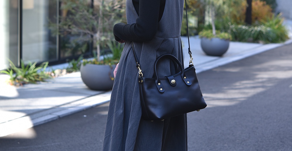 黑色皮革迷你托特包，配有金色金屬配件和可拆卸肩帶，由一位穿著灰色連衣裙的人士展示。