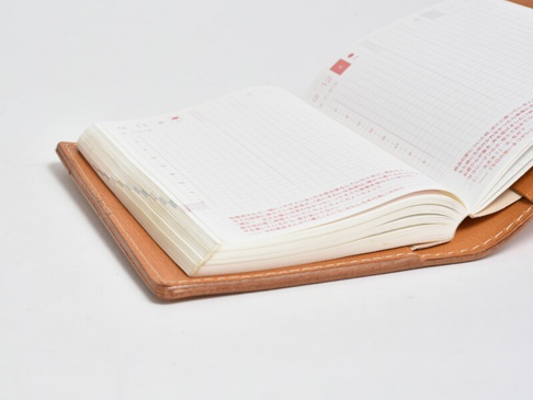 職人手作皮革A6記事本書套，淺棕色皮革，內含一本白色內頁的方格筆記本，筆記本內頁印有紅色字體及時間軸。