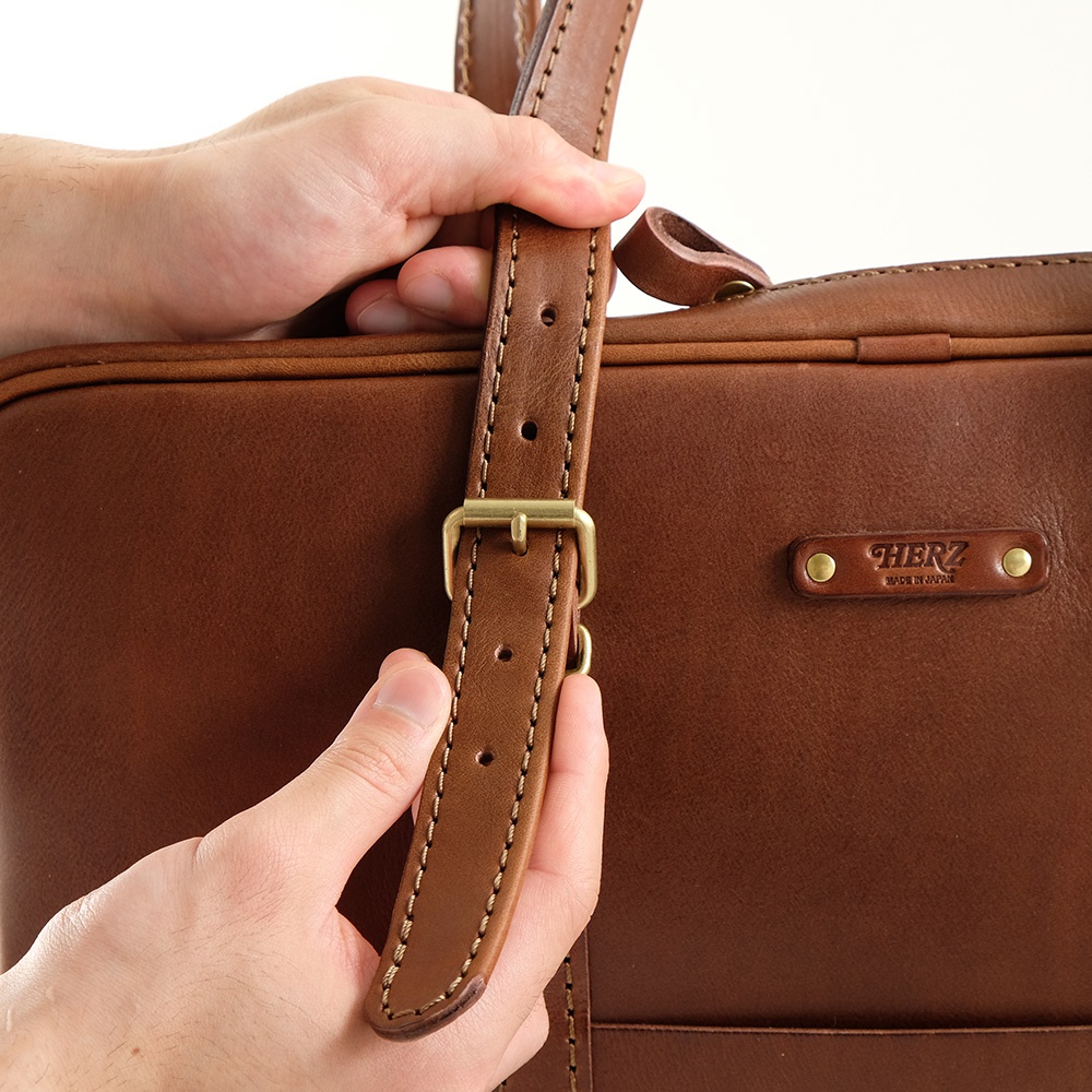職人手作皮革肩背手提兩用公事包，採用質樸的棕色皮革，配有可調節的肩帶和黃銅扣環，包面上印有「HERZ MADE IN JAPAN」的品牌標誌。