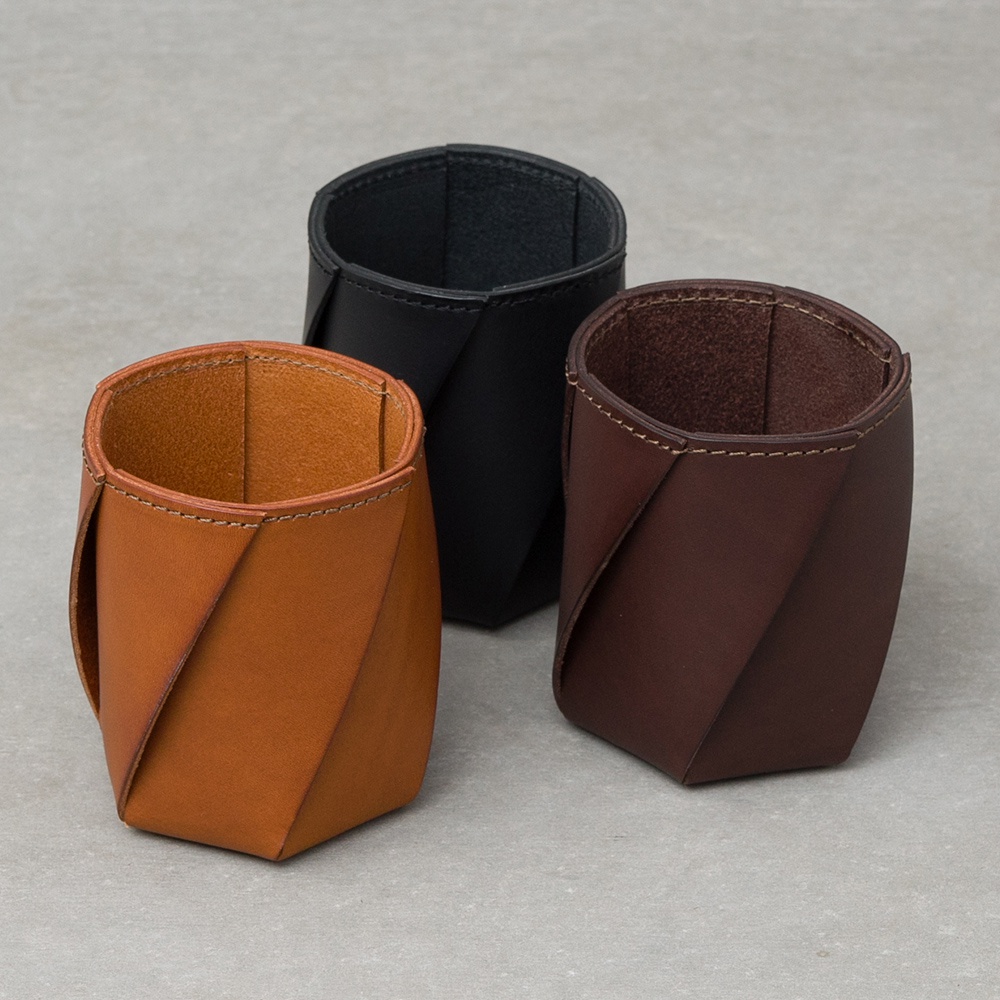 三個皮革收納筆筒，分別為焦糖色、黑色與深棕色，皆為職人手作，呈現六角形扭轉設計，以縫線勾勒出細節。