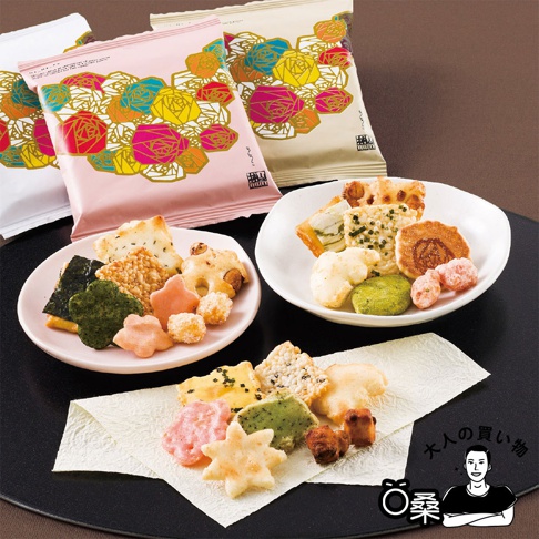這張圖片展示了赤坂柿山的 Nanako 七彩花卉綜合米菓，三包彩色花卉圖案包裝袋擺放在後方，前方兩只盤子與一張襯紙上堆疊了多種造型與口味的日式米菓，包含星星、花朵、海苔片及各類酥脆米餅，右下角配有品牌吉祥物頭像圖示與品牌名稱。