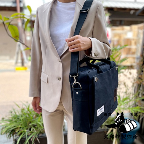 一位身穿米色西裝的女士，左肩斜挎著一個黑色純棉帆布製成的B4公事包，包上配有黑色肩帶及金屬扣環。