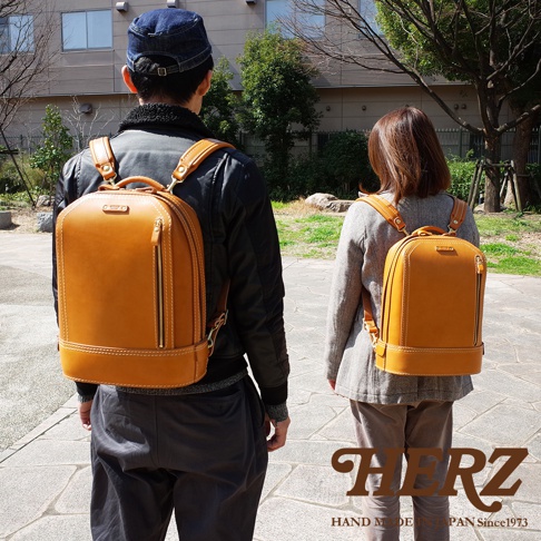 兩位人士身後各背著一個 tan 色皮革立體商務後背包，背包設計簡潔，具有頂部提把和拉鍊開口。