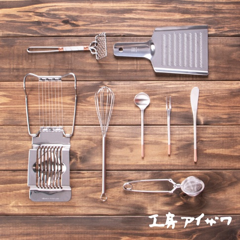 不鏽鋼廚房工具組，包含刨絲器、雞蛋切片器、打蛋器、湯匙、叉子、奶油刀和茶濾勺，皆為金屬材質，部分手柄有玫瑰金飾條，以木紋背景呈現。