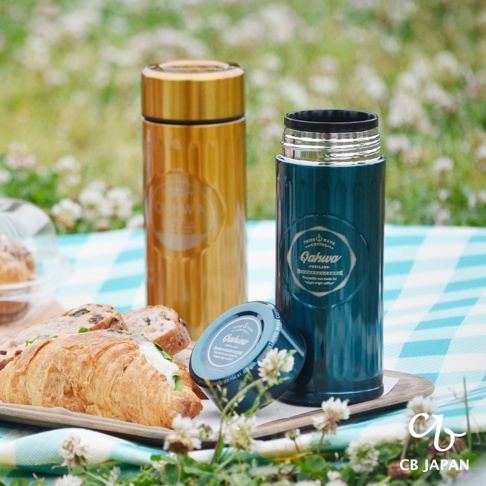 金黃色與藍綠色的城市咖啡保溫杯，均為金屬材質，印有品牌標誌。一旁擺放著可頌麵包與堅果麵包，以及一個藍綠色的杯蓋，整體置於藍白格紋野餐墊上。