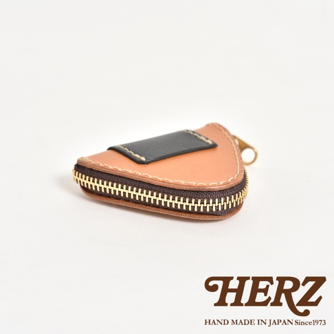 一個日本HERZ職人手作的三角飯糰造型零錢包，選用棕色皮革搭配黑色飾片，邊緣由金屬拉鍊環繞，整體呈現簡約精緻的皮革質感，右下角印有品牌標誌與手工產地說明。