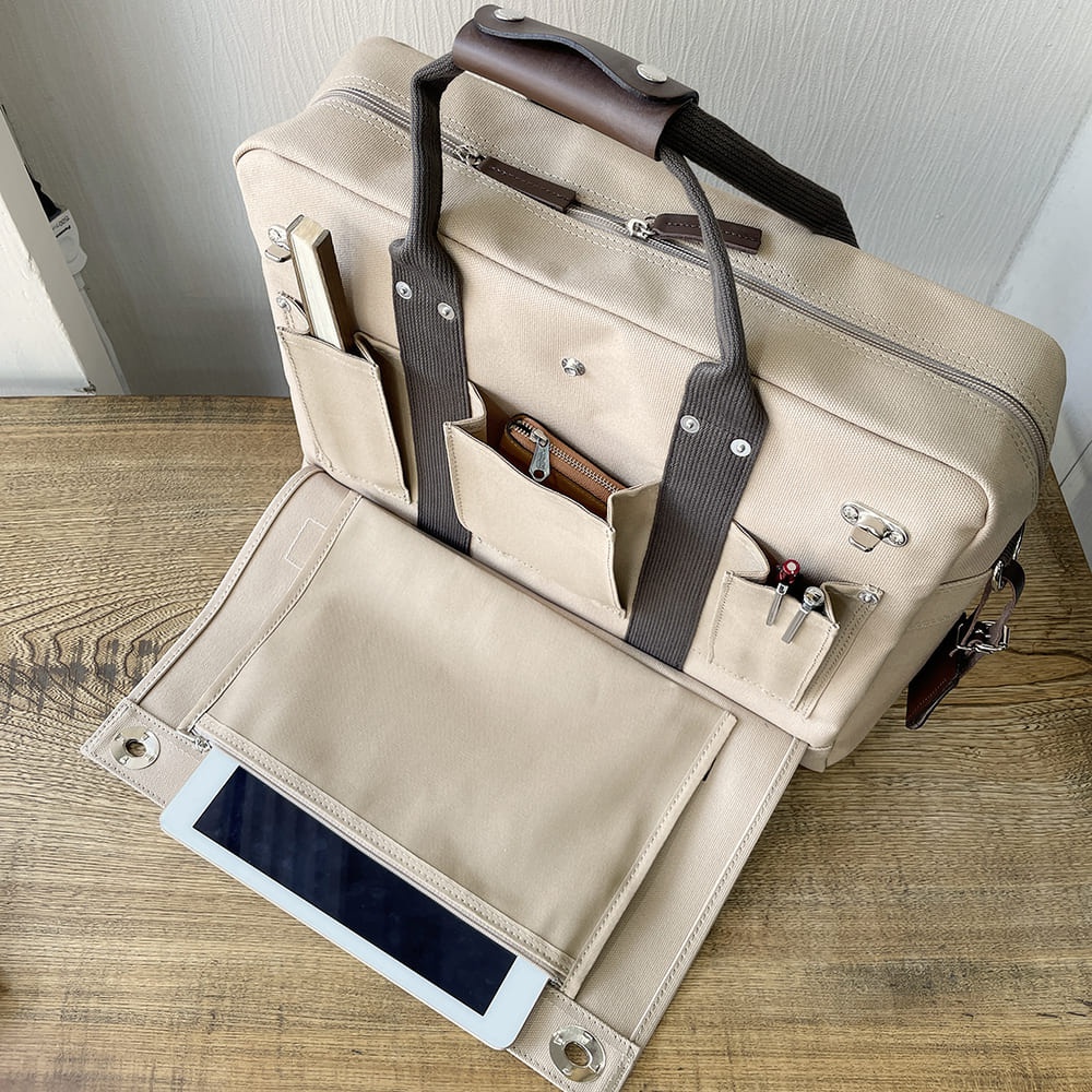 一款米色純綿帆布商務公事包，配有棕色皮革提把和深灰色織帶。包身有多個外部口袋，其中一個口袋內放有棕色皮夾，另一個口袋插有筆。包包前方翻蓋打開，露出一個可放置平板電腦的夾層。