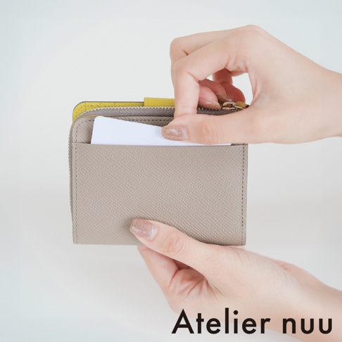 一雙手正拿著一個灰褐色的 Atelier nuu 皮革短夾，其中一隻手從短夾的外層夾層中抽出或插入一張空白的白色卡片，短夾內側邊緣隱約露出亮黃色的細節設計。