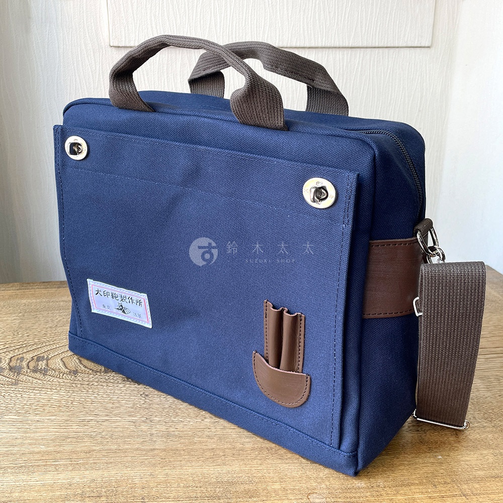藍色純棉帆布三用橫式側背包，附有深棕色皮革裝飾與提把，可拆卸的側背帶，正面有兩個金屬鈕扣，一個標籤和一個皮革筆插。