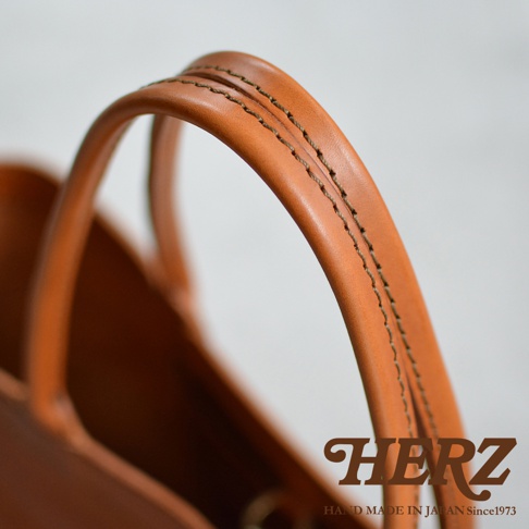 特寫鏡頭展示了職人手作皮革托特包的提把，以細膩的縫線工藝呈現，包身為溫潤的焦糖棕色皮革。