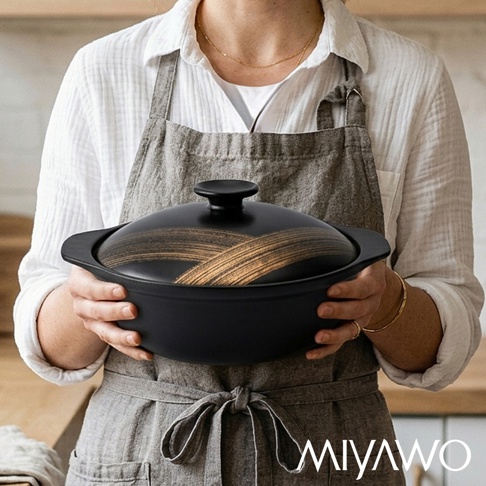 一位穿著白襯衫與灰色圍裙的人雙手捧著一只黑色MIYAWO日本宮尾直火系列陶土湯鍋，鍋蓋中央繪有金色的交叉筆觸紋路，鍋身設計簡約現代，呈現出質感優雅的廚房料理氛圍。