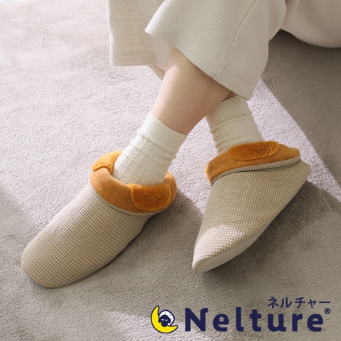 一雙米色網格紋的睡眠專用保暖腳踝室內拖鞋，鞋口處有橘色絨毛裝飾，由穿著白色襪子的人穿著，背景是灰色地毯。