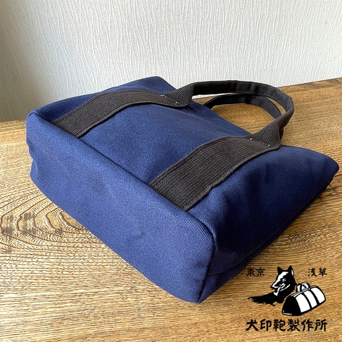 深藍色帆布水桶托特包，配有黑色織帶提把，包身底部和側面可見，整體造型簡潔。