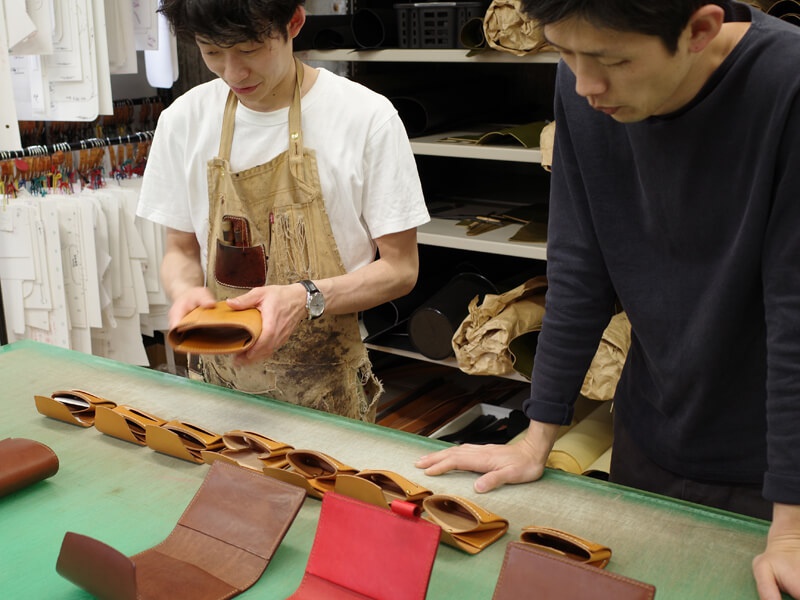 兩名男性在綠色工作檯前，一名男子身穿圍裙，手持一個棕褐色皮革 A6 記事本書套。工作檯上擺放著多個相同款式的記事本書套，以及其他皮革製品，如紅色、棕褐色的筆記本封面。背景可見架子上擺放著皮革材料和工具。