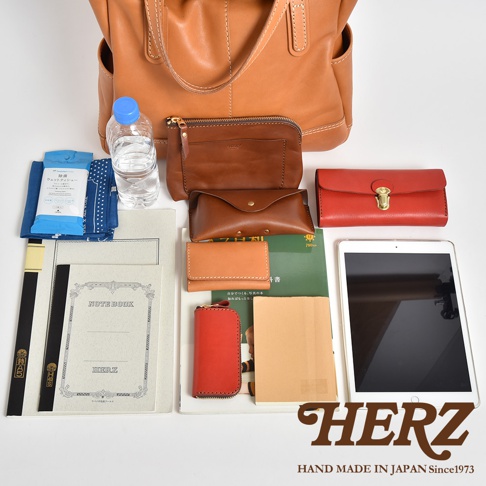 俯視視角展示了日本 HERZ 職人手作皮革托特包及其收納內容物，包含透明塑膠水瓶、濕紙巾、藍色布料、筆記本、長夾、多個皮革小包與收納袋、記事本以及一台平板電腦，整體呈現出豐富且有質感的日式皮革手工藝風格。