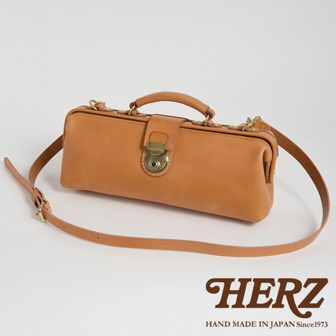 HERZ 品牌標誌，下方印有 HAND MADE IN JAPAN Since1973，搭配一個展示於純白背景上的棕色皮革橫長形包款，包款設有提把、金色鎖扣和可調式長肩帶。