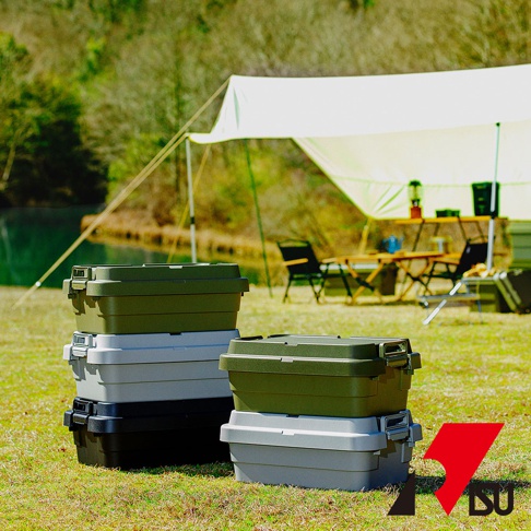 草地上堆疊著三款 TRUNK CARGO 可堆疊多功能耐重收納箱，分別為綠色、灰色與黑色。收納箱為塑膠材質，帶有銀色金屬扣環。背景是露營場景，有天幕、桌椅和湖泊。