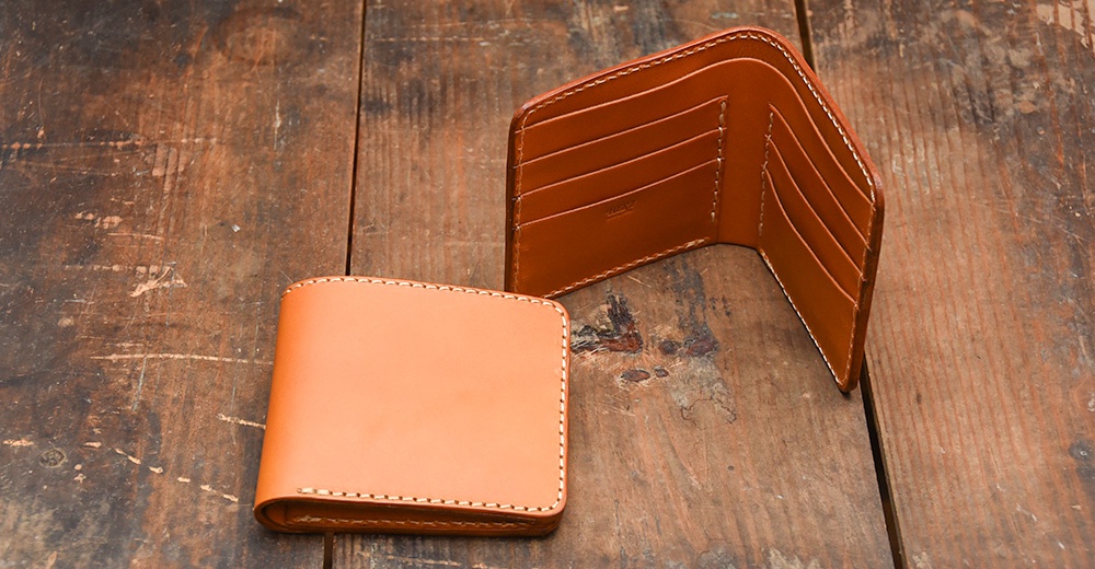 職人手作皮革二折橫式卡片紙鈔小短夾，呈現自然棕色調，光滑皮革表面。一款錢夾閉合，另一款錢夾攤開，露出多個卡槽，所有邊緣皆以白色車線縫合。