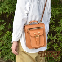 職人手作的棕色皮革立體口袋兩用單肩包，正面有拉鍊口袋、翻蓋口袋和金屬釦帶，頂部有提把，搭配細長肩帶。