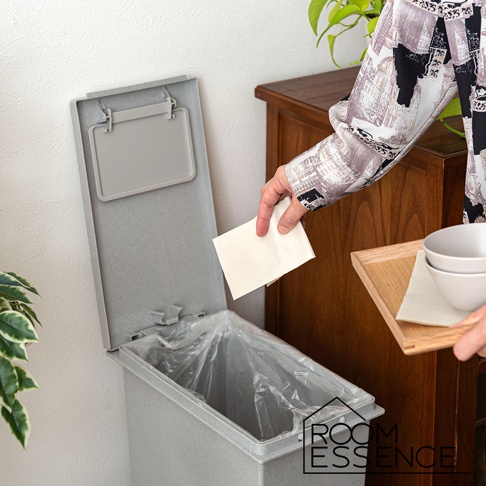 一個灰色、附蓋的簡約垃圾桶，頂部有可開合的蓋子，內部裝有透明垃圾袋。一位女士正將紙巾丟入垃圾桶，手中拿著一個木製托盤，托盤上有兩個白色碗。