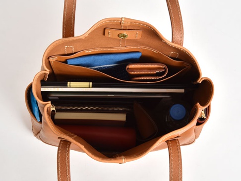 職人手作皮革直式肩背托特包，呈現自然棕色調，包內整齊收納著書籍、筆記本、錢包、藍色織物及一瓶水，展現其寬敞且分層的實用性。