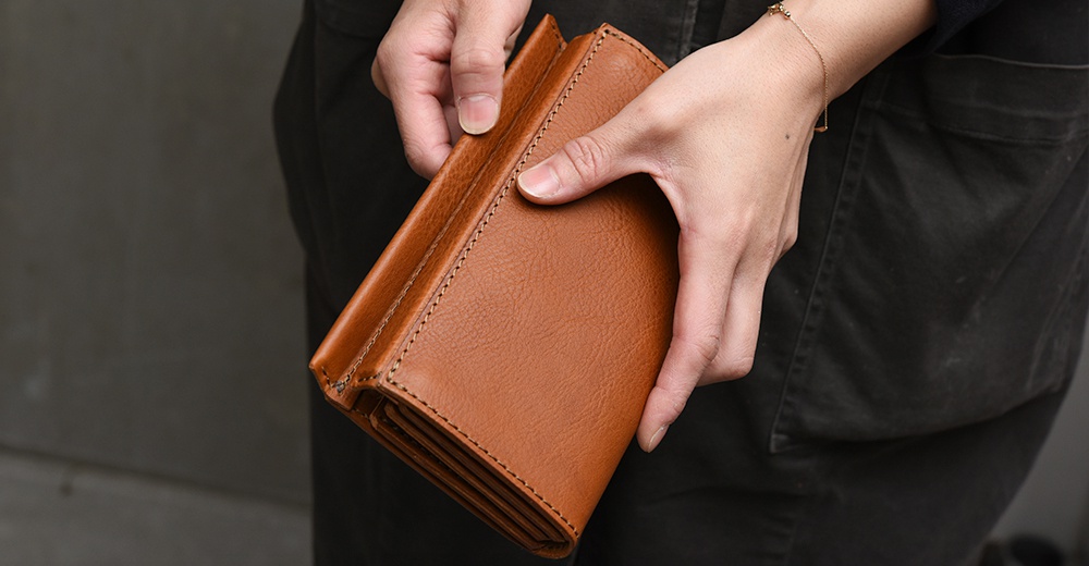 一位穿著深色工作服的人雙手捧著一個 HERZ 日本職人手作的棕色皮革長夾，長夾表面呈現自然的皮革紋理，邊緣縫線工整細緻，展現出簡約質感與扎實的手工工藝。