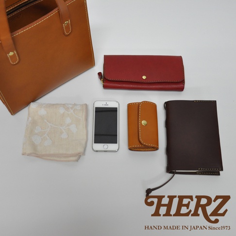 職人手作皮革直式托特包 T-44 XS，展示了咖啡色皮革托特包、紅色長夾、白色手機、咖啡色鑰匙包，以及一塊米白色印花方巾。