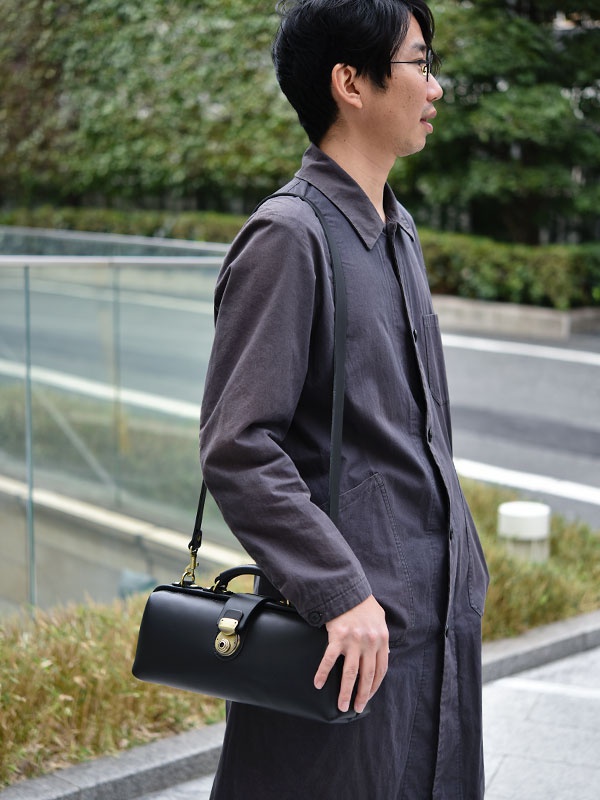 黑色皮革橫長形杜勒斯單肩包，配有金色鎖扣和提把，由一位身穿深灰色長外套的男士斜背著。