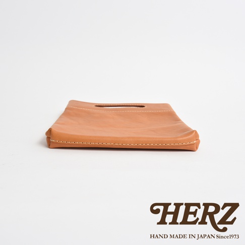 一款日本HERZ手工製作的棕色皮革收納袋，從低角度仰拍呈現其簡約的扁平長方形外觀，袋口上方設有挖空的提把設計，邊緣飾有細緻的淺色縫線，右下角印有品牌名稱與創立年份標誌。