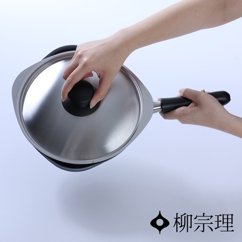 雙手握持柳宗理品牌的雙網紋單手鐵鍋，金屬質感鍋蓋與黑色手柄對比鮮明，整體呈現極簡現代風格，右下方印有柳宗理品牌名稱與其經典標誌。
