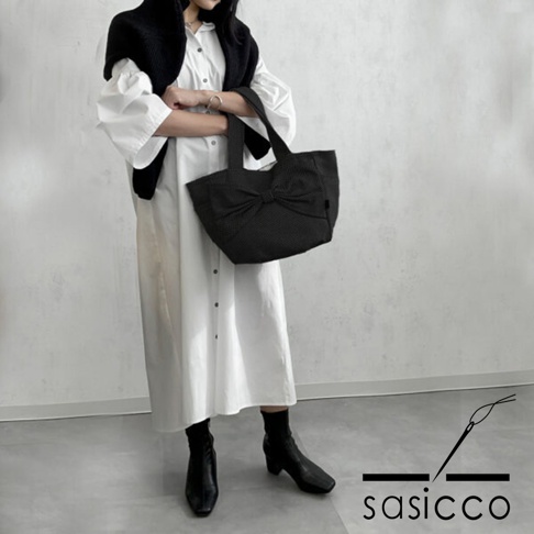 以黑色三河木綿刺子織製成的蝴蝶結托特包，提把設計，包身有精緻的織紋，由一位身穿白色長洋裝、肩披黑色毛衣的女性手持。