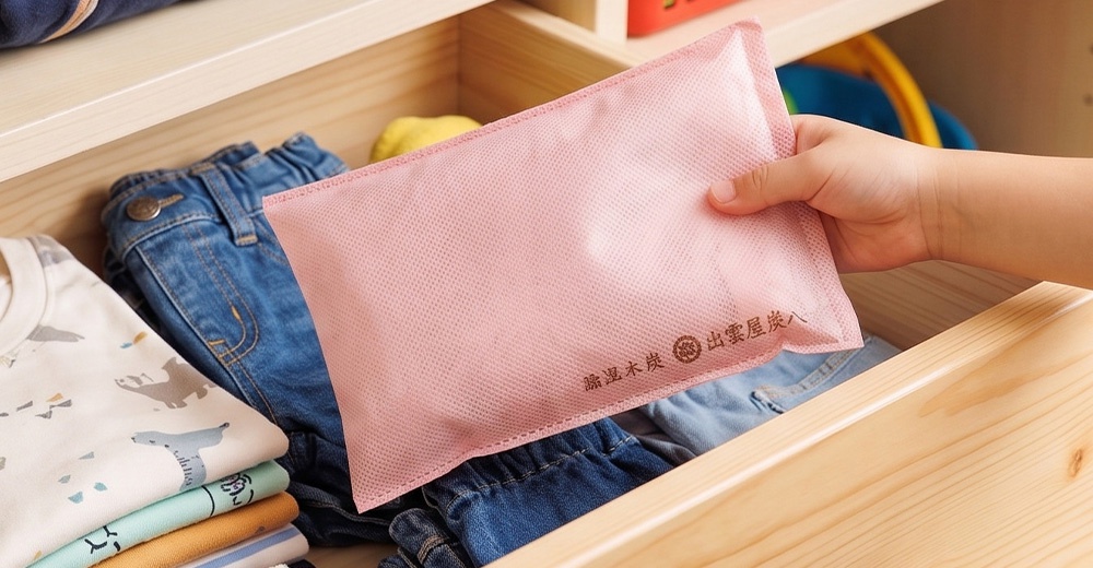 一隻手正將一個淺粉色、透氣網狀材質的長方形除濕包放入木質抽屜中，抽屜內同時放置著摺疊整齊的衣物與牛仔褲，除濕包上印有深色品牌文字。