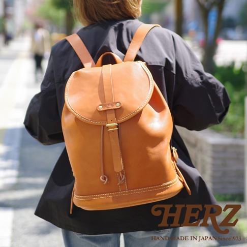 一個棕色皮革後背包，有單扣束口設計，肩帶和包身皆為皮革材質，包底有白色縫線，由一人背著。