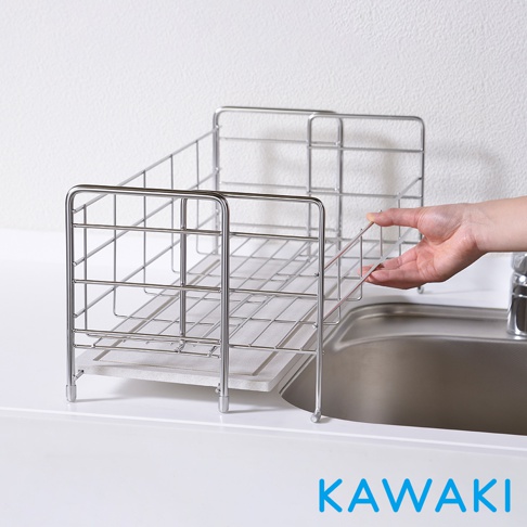 銀色不鏽鋼線製成的伸縮式碗盤瀝水架，設有網格層架，底部有淺灰色珪藻土吸水墊，架子置於廚房水槽旁，一隻手正在調整架子。
