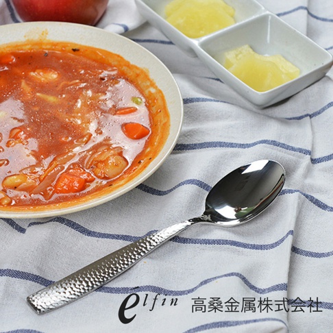 日製錘目紋不鏽鋼餐匙，銀色，光滑的湯匙頭，帶有錘擊紋理的餐匙柄，放在藍白條紋布上，旁有一碗湯和兩個小碟。