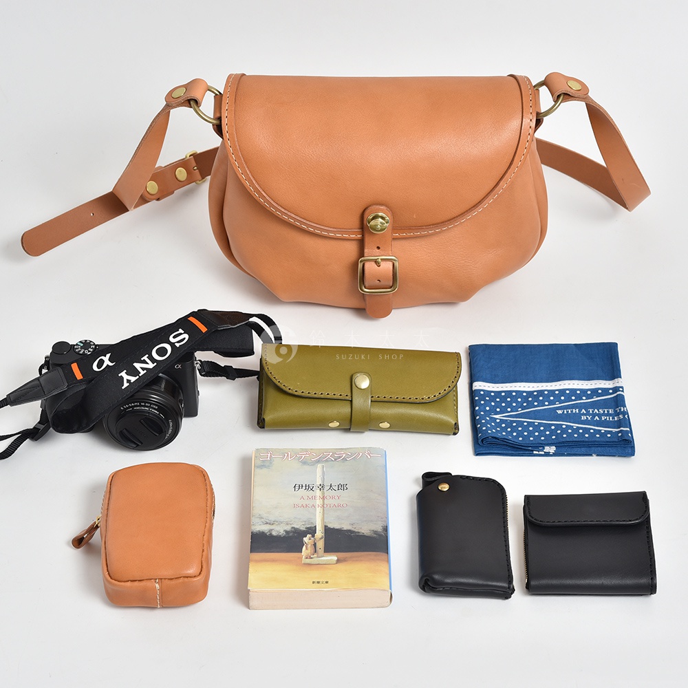 日本HERZ皮革休閒旅行馬鞍包，搭配黑色相機、綠色皮夾、藍色手帕、棕色小皮夾和日文書籍，皆擺放在白色背景上。