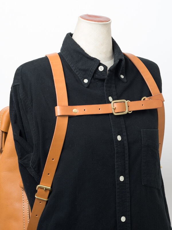 職人手作皮革背包用胸前扣帶，寬 2.4 公分，採用淺棕色皮革，搭配黃銅色金屬釦環，可調節長度，固定於黑色襯衫上。
