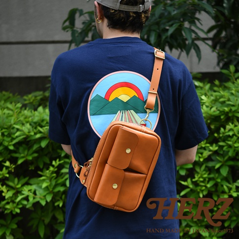 一款皮革斜背包，採用棕褐色皮革製成，設有兩個帶有金屬鈕扣的外部口袋，以及一個拉鍊主隔層。背包帶有可調節的肩帶，通過金色金屬扣連接。