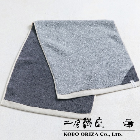 深灰色與淺灰色棉麻混紡毛巾，兩色拼接設計，觸感柔軟，邊緣有白色滾邊，一角有品牌標籤。