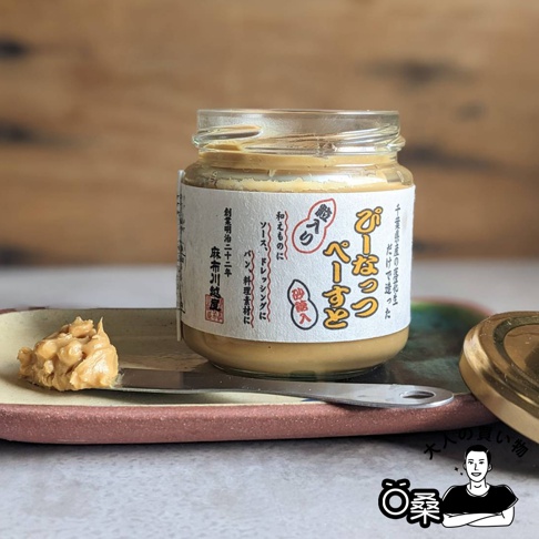 玻璃罐裝的顆粒花生醬，醬體呈淺棕色，上面貼有日文標籤，標籤上有紅色和白色的字體。一小塊花生醬被塗抹在陶製的淺盤上，旁邊放著一把金屬抹刀。