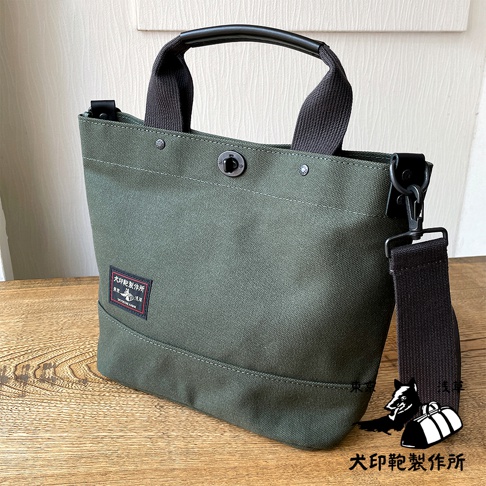 軍綠色帆布材質的兩用托特包，配有黑色皮革提把和可拆卸肩帶，包身正面有紅色標籤，標有日文文字和黑犬圖案。