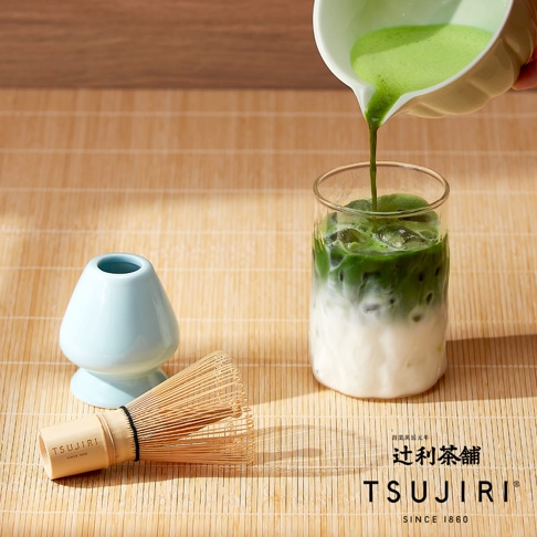 辻利茶舖的抹茶製作場景，在竹製編織底板上，左側擺放著淡藍色陶瓷茶筅座與一支竹製茶筅，右側則有手持淺綠色片口碗將鮮綠色抹茶液倒入裝有冰塊與牛奶的玻璃杯中，呈現抹茶與牛奶層次分明的質感。